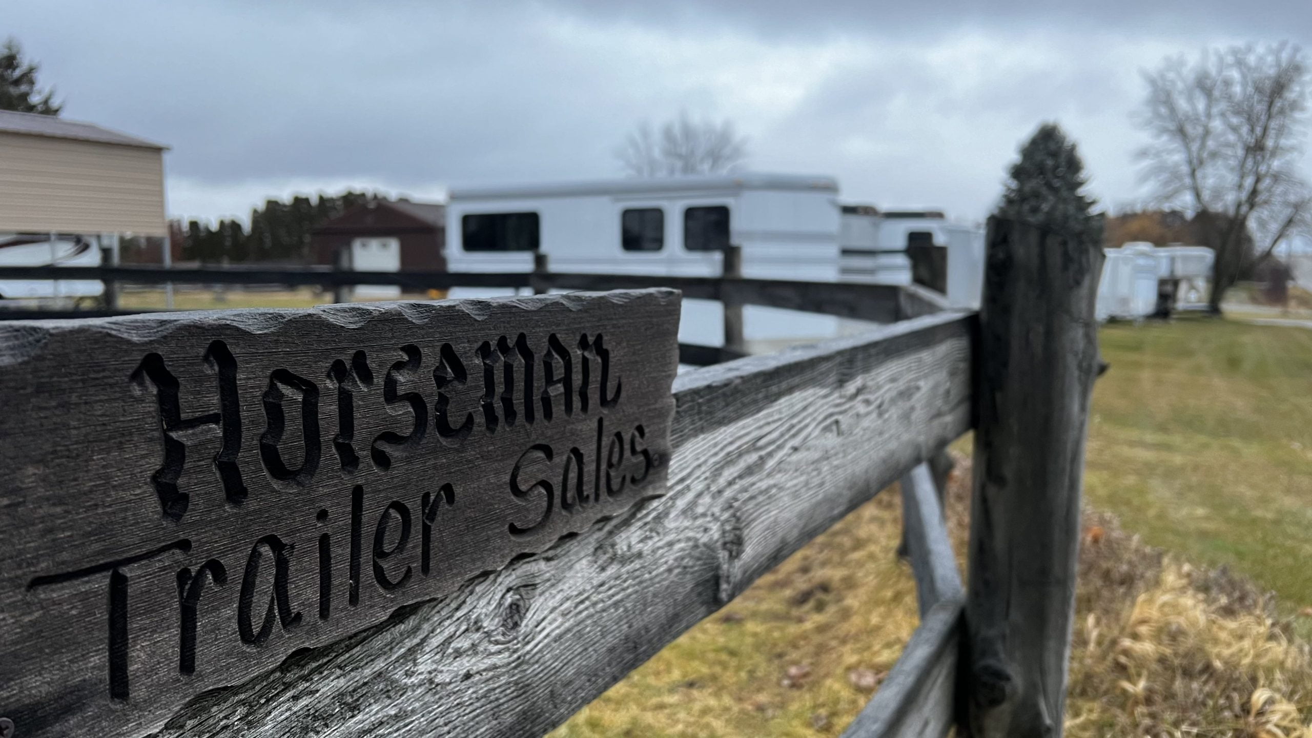 a small wooden sign on the fence of a round pen with a line of horse trailers in the distance.